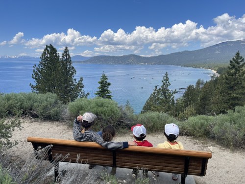 A family sits on a bench overlooking a scenic lake and mountains under a blue sky with fluffy clouds.