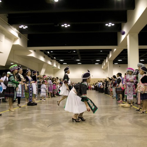 A group of dancers in traditional attire perform in a large indoor space, with spectators watching from the sides.