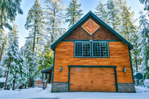 A cozy wooden cabin surrounded by snow-covered trees, featuring large windows and a stone-accented garage door.