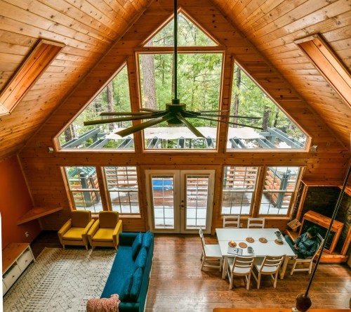 Cozy wooden cabin interior with large windows, a dining table, and a living area featuring a blue sofa and yellow chairs.