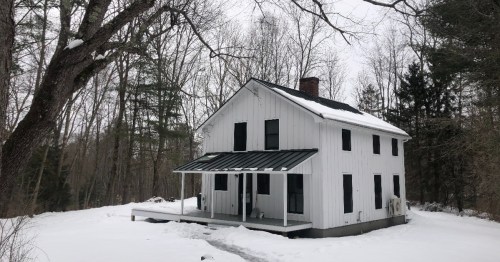 A white two-story house with a porch, surrounded by snow and bare trees in a winter landscape.