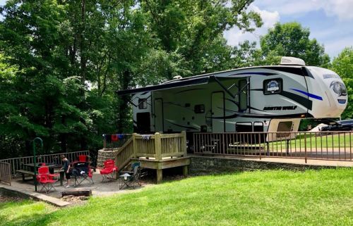 A parked RV on a wooden deck surrounded by trees, with red chairs and a fire pit nearby.