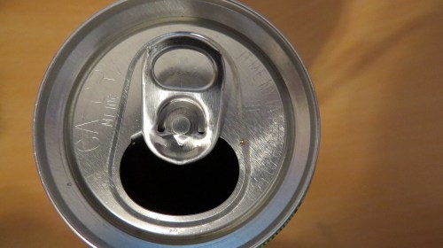 Close-up of the top of a soda can, showing the pull tab and opening against a wooden surface.