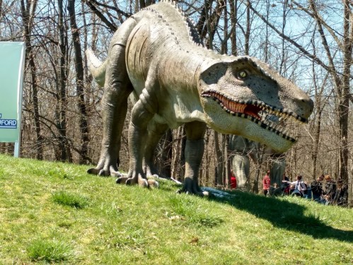 A large dinosaur sculpture stands on a grassy hill, surrounded by trees and people in the background.