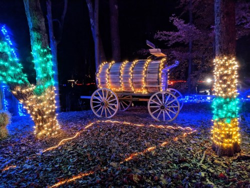 A wooden wagon decorated with colorful lights, surrounded by illuminated trees in a dark forest setting.
