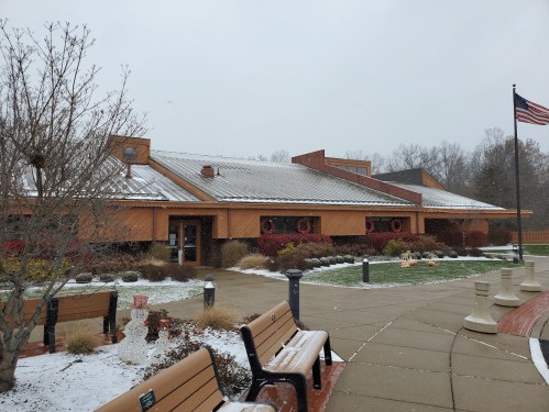 A snowy scene of a modern building with a metal roof, surrounded by benches and festive decorations. An American flag flies.