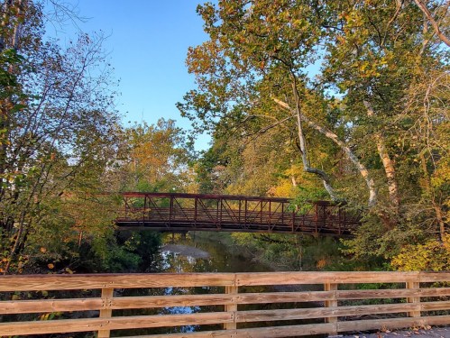 A wooden bridge spans a calm river, surrounded by trees with autumn foliage under a clear blue sky.