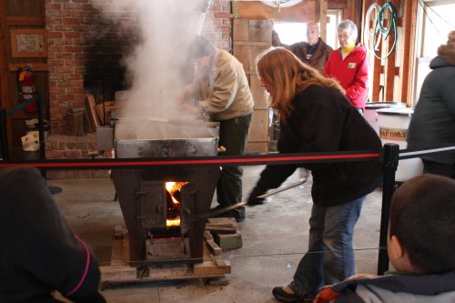 A blacksmith works at a forge, with smoke rising, while onlookers watch in a rustic workshop setting.
