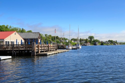 A peaceful waterfront scene with boats docked, a wooden pier, and lush greenery under a clear blue sky.