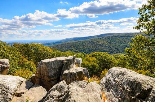 Scenic view of rocky outcrops overlooking a lush green valley and rolling hills under a blue sky with fluffy clouds.