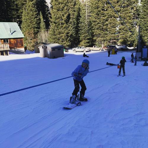A child snowboarding on a snowy slope with trees and buildings in the background. Other kids are nearby.
