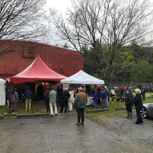 A gathering of people under tents outside a red building, surrounded by trees and a grassy area.