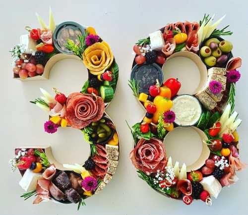 A colorful charcuterie board shaped like the numbers 3 and 8, adorned with meats, cheeses, fruits, and flowers.