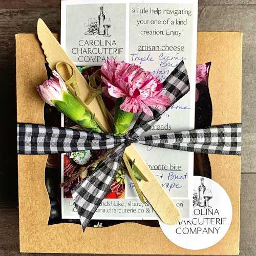 A gift box from Carolina Charcuterie Company, decorated with flowers and a wooden spreader, featuring a cheese guide.