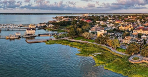 Aerial view of a coastal town with waterfront, boats, and lush greenery under a cloudy sky.