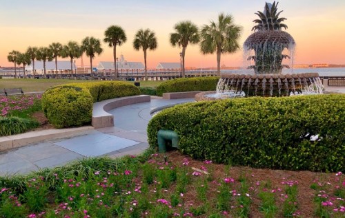 A fountain surrounded by lush greenery and flowers, with palm trees and a sunset in the background.