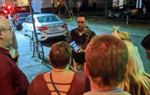 A group of people gathered outdoors, listening to a man holding a phone, with cars and bicycles in the background.
