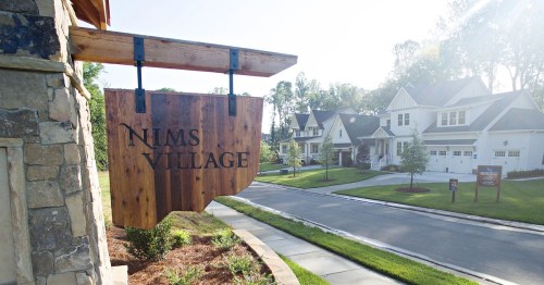 Sign for Nims Village with a residential neighborhood in the background, featuring modern homes and a sunny atmosphere.