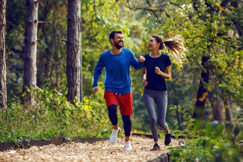 A man and woman jog together on a forest path, smiling and enjoying their run amidst lush greenery.