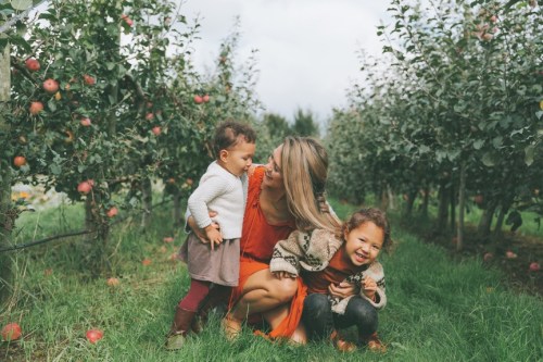 A woman crouches in an apple orchard with two young girls, surrounded by apple trees and green grass.