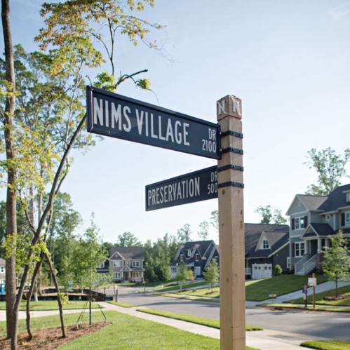 Street sign at a neighborhood intersection, labeled "Nims Village Dr" and "Preservation Dr," with houses in the background.