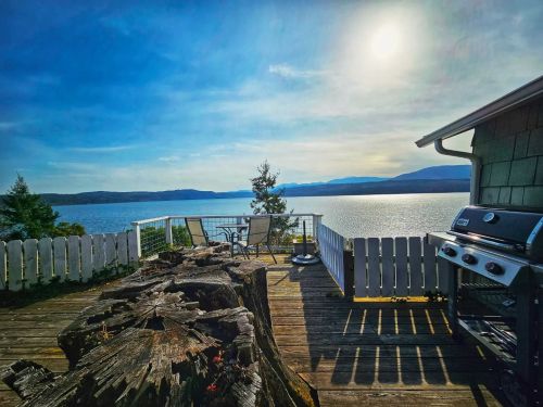 Scenic view of a lake from a deck with a grill, chairs, and a wooden stump under a bright blue sky.