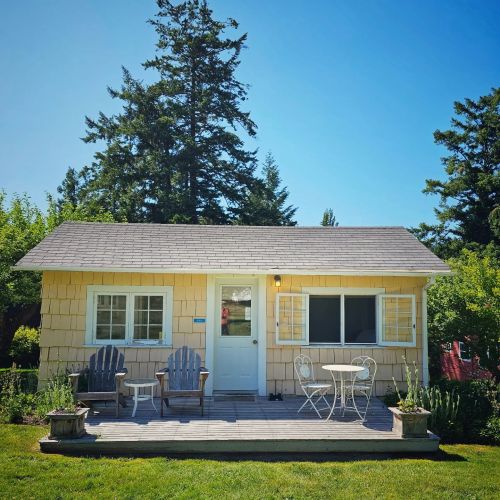 A charming yellow cottage with a wooden deck, two chairs, and a small table, surrounded by greenery and trees.