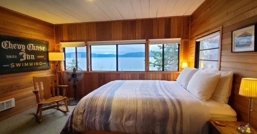 Cozy wooden bedroom with a large bed, rocking chair, fan, and windows showcasing a scenic lake view.