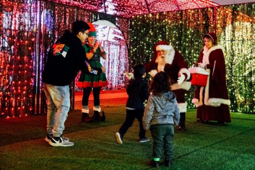 A festive scene with Santa, two elves, and children interacting in front of colorful holiday lights.