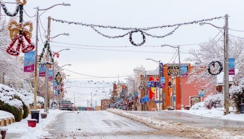 Snow-covered street decorated with holiday wreaths and lights, lined with shops and trees in a winter landscape.