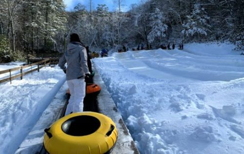 A person in winter gear walks towards a snow-covered tubing slope, with others in the background enjoying the snow.
