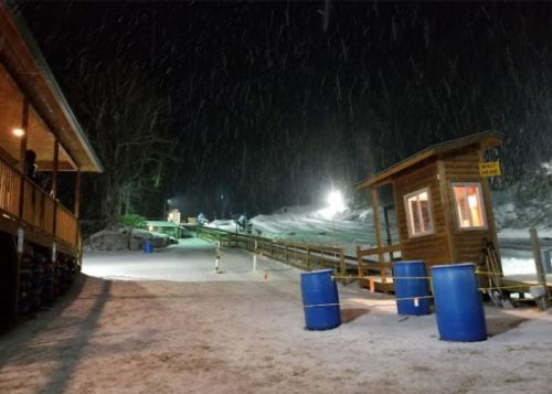 A snowy scene at a ski resort with a wooden building, a ticket booth, and illuminated slopes in the background.