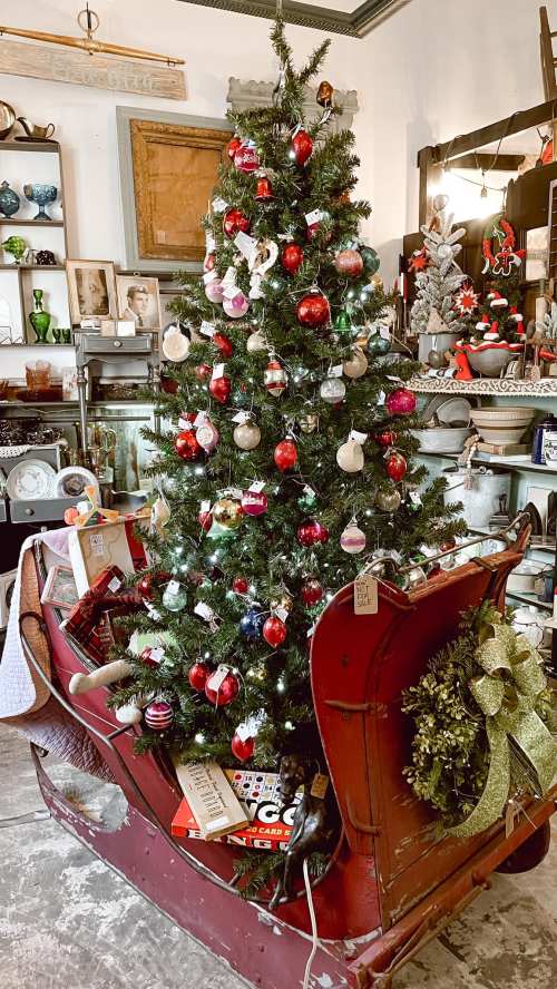 A decorated Christmas tree in a vintage sleigh, surrounded by ornaments and festive decor in a cozy shop.