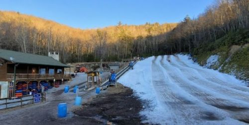 A snow tubing hill with multiple lanes, surrounded by trees and a lodge in the background, on a sunny winter day.