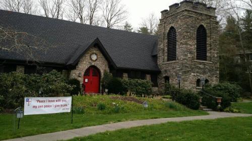 A stone church with a red door, surrounded by greenery and a sign that reads, "Peace I leave with you..."