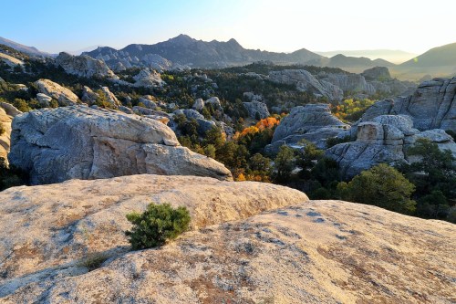 A scenic view of rocky terrain and distant mountains, with autumn foliage in the valley under a golden sunset.