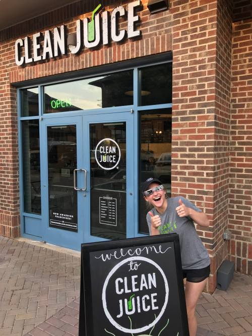 A smiling person gives a thumbs up in front of a Clean Juice store with a welcome sign.
