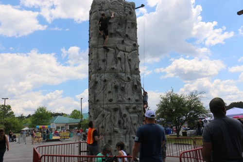 Two climbers ascend a rock wall at an outdoor event, with spectators watching nearby under a partly cloudy sky.