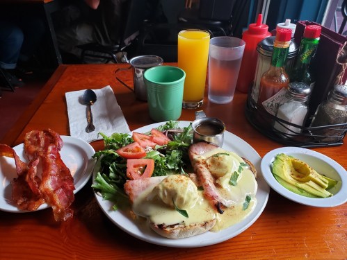 A plate of eggs Benedict with bacon, salad, and tomatoes, accompanied by a glass of orange juice and condiments.