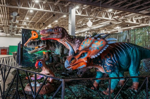 Colorful, life-sized dinosaur models displayed in a museum-like setting with a forest backdrop.