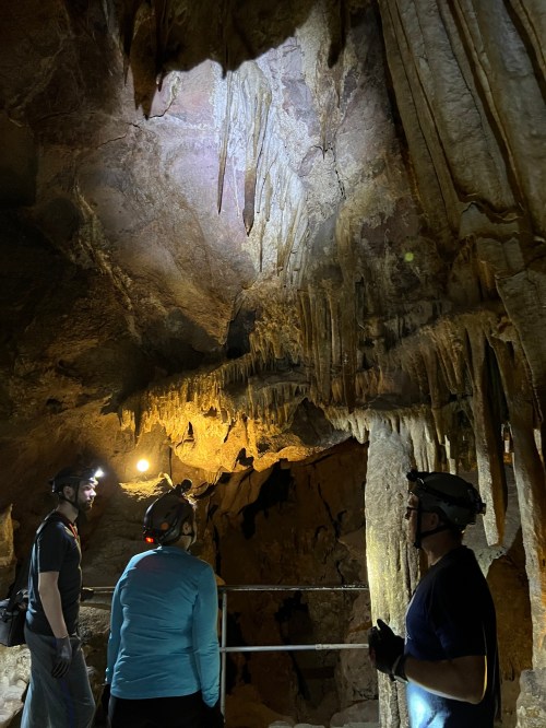 Three people in helmets explore a cave, illuminated by headlamps, with stalactites hanging from the ceiling.