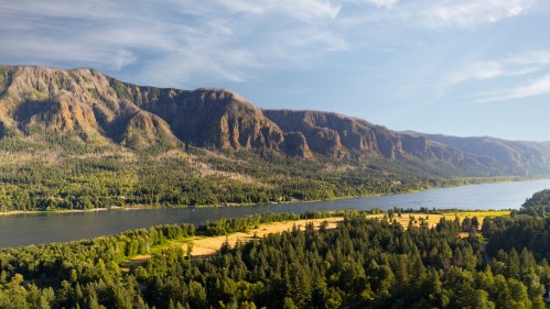 A scenic view of mountains and a river, surrounded by lush greenery and fields under a clear blue sky.