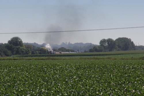 A train travels through a green field, with smoke rising in the background against a clear blue sky.