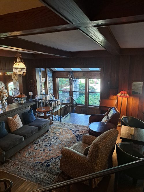 Cozy living room with wooden walls, a large window, a sofa, armchairs, and decorative rugs. Warm, inviting atmosphere.