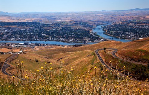 A scenic view of rolling hills, a winding river, and a town nestled in a valley under a clear blue sky.