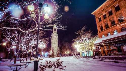 Snow-covered plaza at night, featuring a clock tower and trees adorned with twinkling lights.