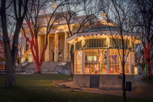 A festive gazebo adorned with lights, surrounded by trees, near a historic building illuminated for the holidays.