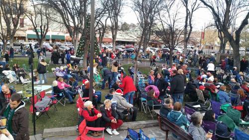 A crowded outdoor event with people sitting in chairs, some wearing festive attire, surrounded by trees and buildings.