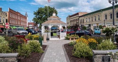 A charming town square with a gazebo, flower beds, and historic buildings under a blue sky with scattered clouds.