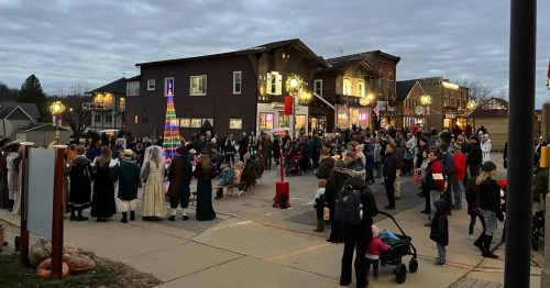 A crowd gathers in a festive town square, with a colorful Christmas tree and holiday decorations under a cloudy sky.